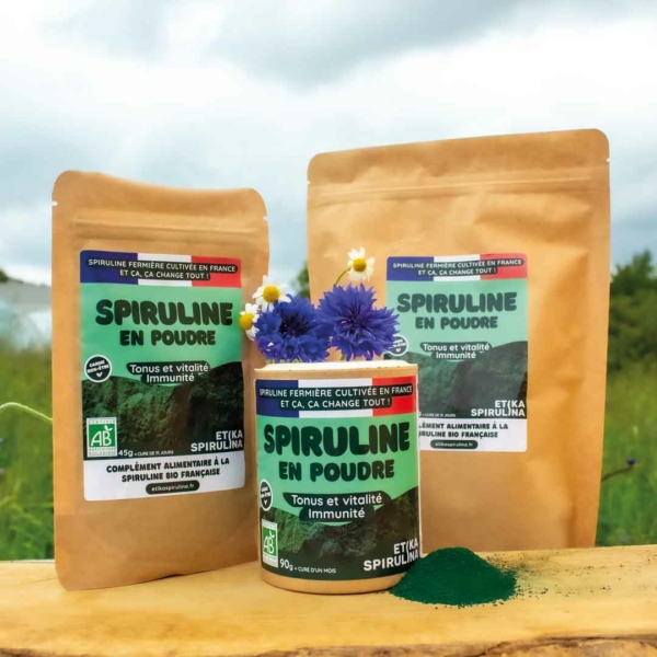 Mise en situation des tailles de spiruline en poudre avec nos serres de spiruline et un champ de fleurs en arrière-plan